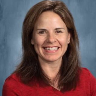 Jill Hoort smiles toward the camera against a neutral bluish background. Hoort wears a red blouse. Hoort has brown shoulder-length hair.