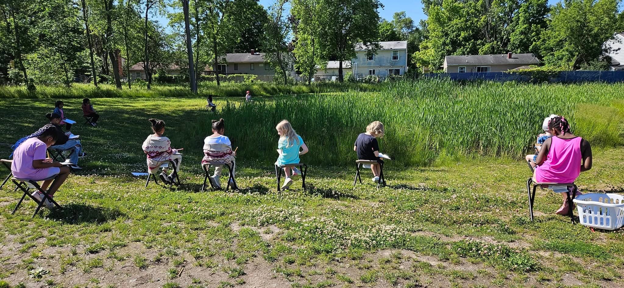 A group of students sit on chairs in a grassy area and take notes while observing a pond outside their school.