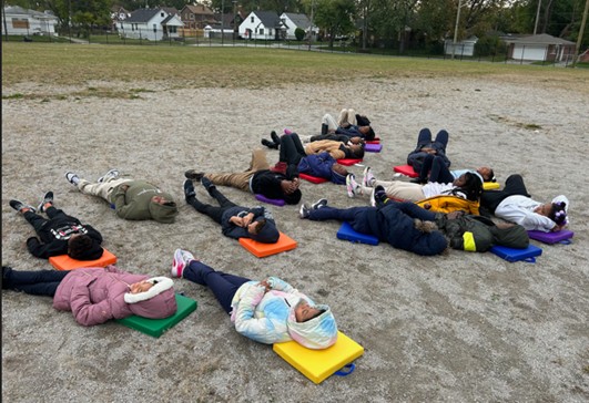 A group of students lay on the ground and gaze upward toward the sky. The students rest on colorful pillows and wear winter coats in various colors and designs. 