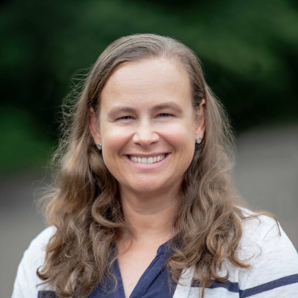 Jenny Sanford smiles for the camera in an outdoor setting. Sanford wears a navy blue blouse under a gray cardigan with a navy blue stripe on it. Sanford's hair is dark blonde and wavy and falls past the shoulders.