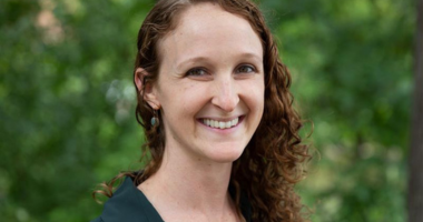 Alex Allweiss poses for a photo, smiling in front of green foliage on MSU's campus. Alex's auburn hair is curly and falls past the shoulders.