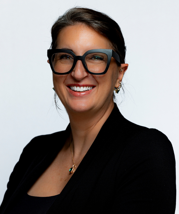 Sarah Gretter Atkins smiles for the camera against a neutral white backdrop. Gretter Atkins wears a black shirt under a black blazer and black-rimmed glasses. Gretter Atkins has several earrings and a gold necklace with a green pendant. Gretter Atkins has dark hair, pulled back and away from the face.