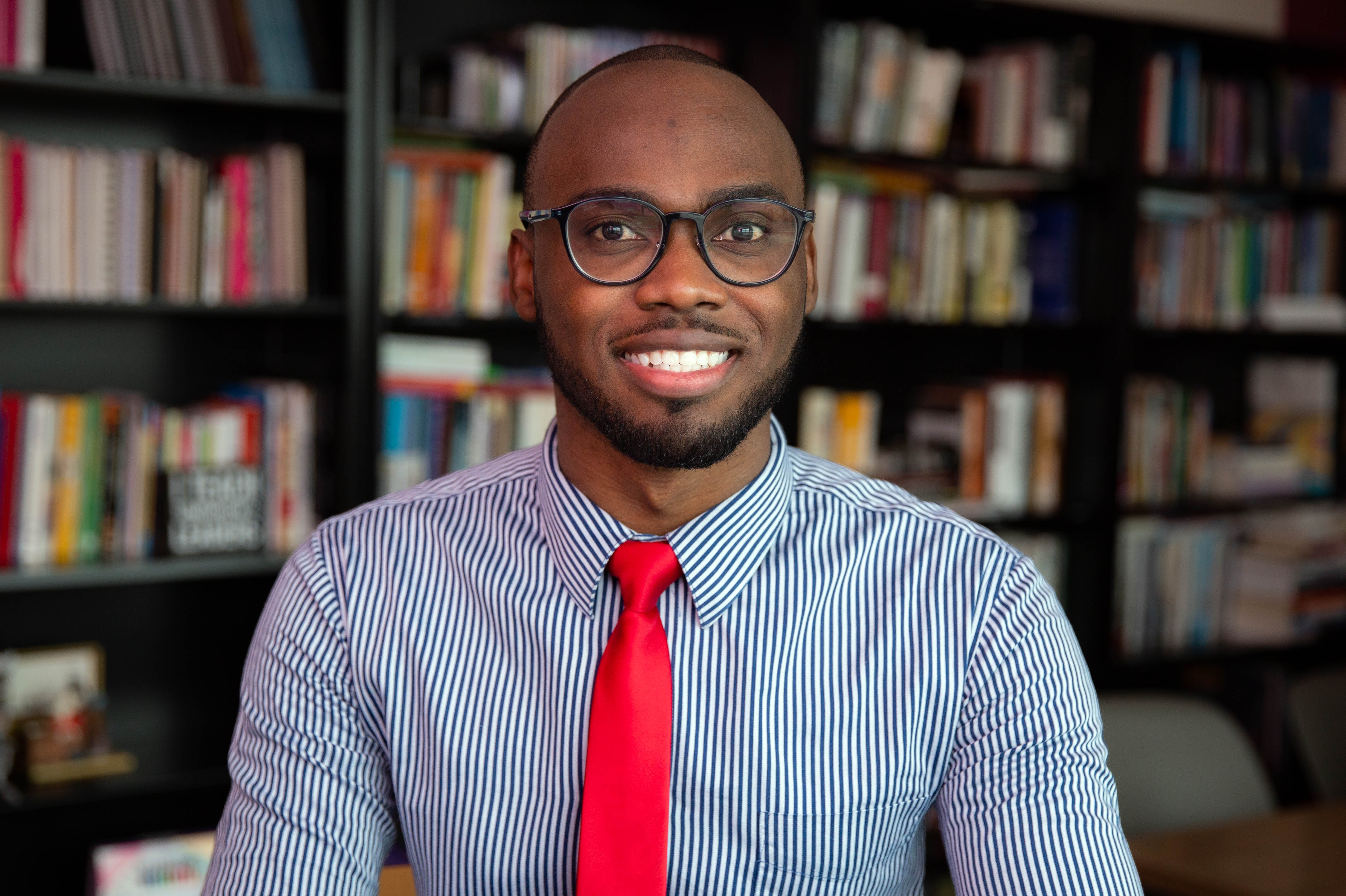 Jermaine Howell headshot. Jermaine poses for the camera against a full bookshelf. Jermaine wears a blue button-down shirt, a bright red tie and dark-rimmed glasses. Jermaine has black hair and beard, both are cut close to the face and head.