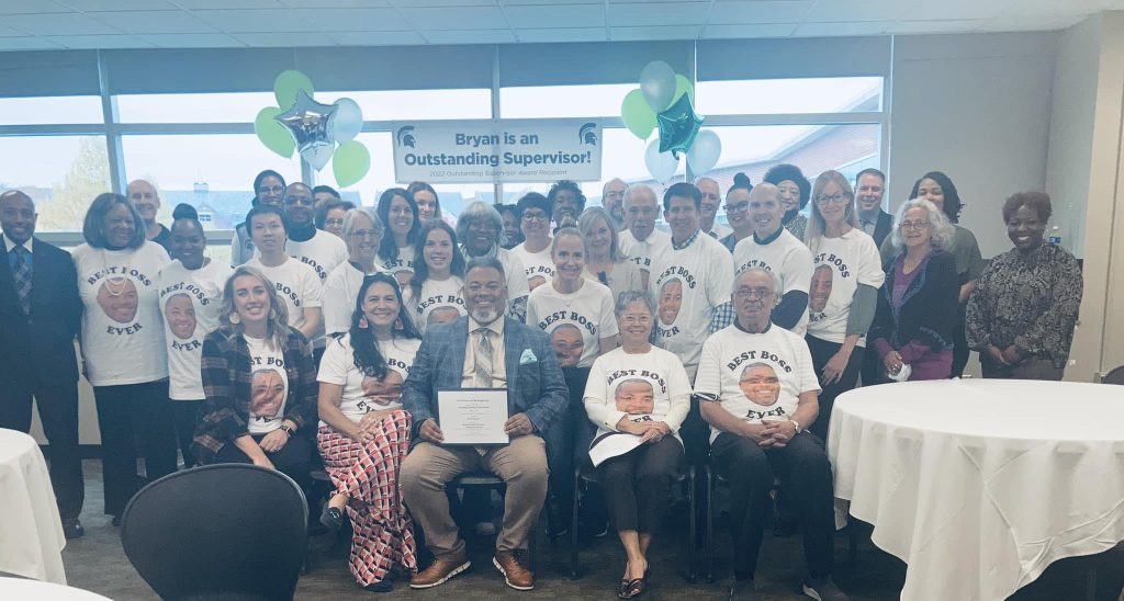 Bryan Beverly sits among a large group of friends and colleagues during a celebration for Outstanding Supervisor. Most attendees wear shirts with Beverly's face on it and the words 