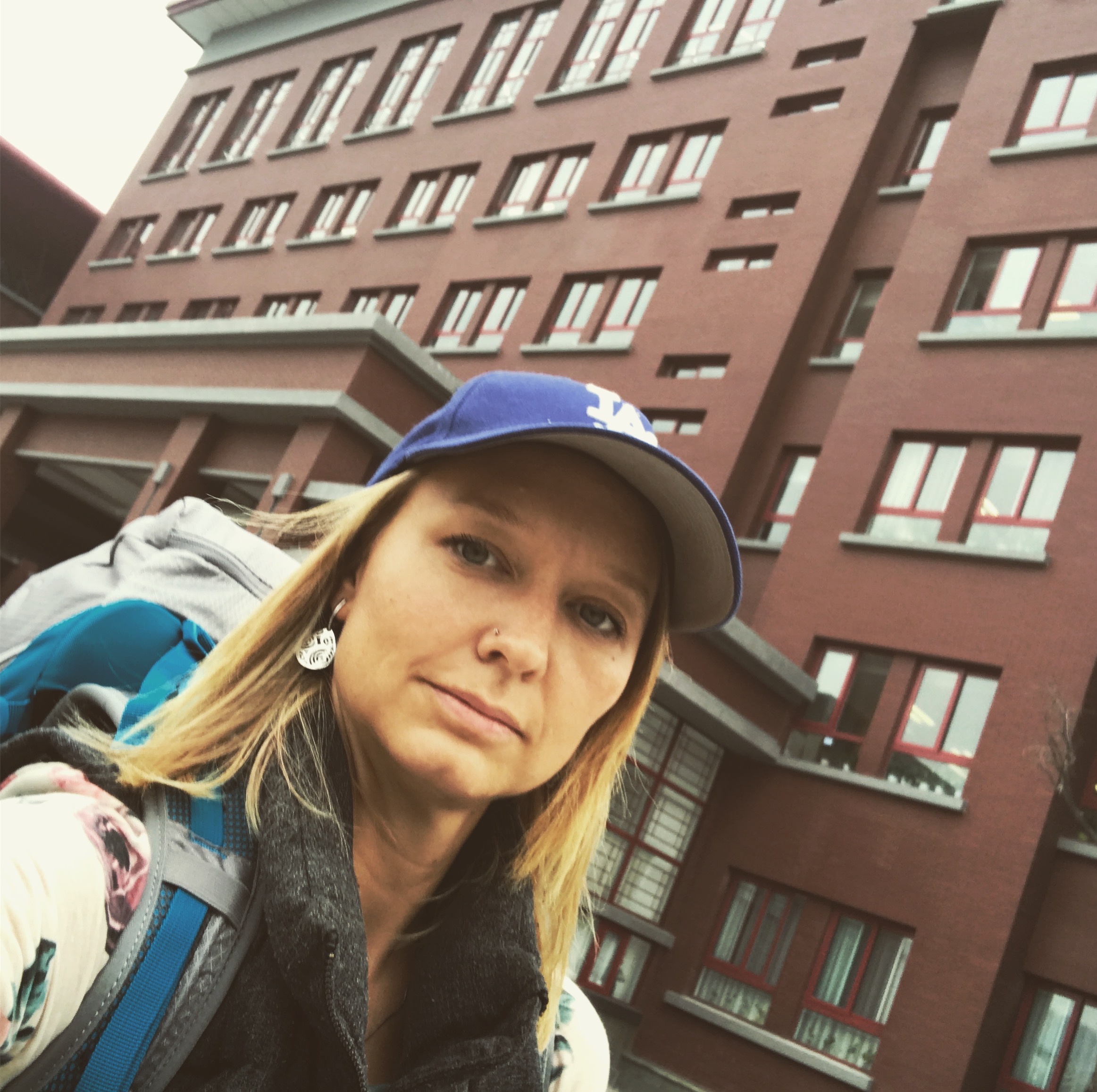 Elicia Bullock takes a photo while walking outside in an industrial area. A dark red, multi-story brick building is behind Elicia. Elicia wears a blue ball-cap over Elicia's long and straight blonde hair.