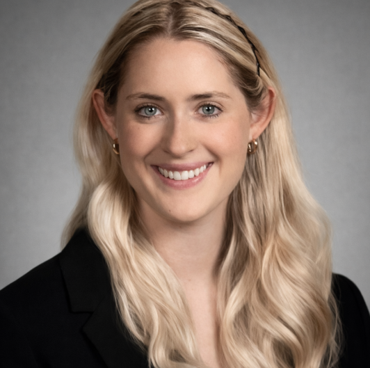Clare Kinna smiles for the camera against a neutral gray backdrop. Kinna wears a black blouse. Kinna's hair is blonde and falls past the shoulders.