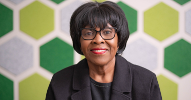 Patricia Edwards poses for a photo against a hexagon backdrop of various shades of green and gray. Edwards wears a black blouse and blazer. Edwards' black hair is short and chin-length. Edwards wears black-rimmed glasses.