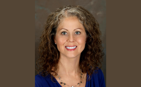 Elizabeth Hicks smiles for the camera against a neutral brown background. Hicks wears a royal blue top and layered silver necklaces. Hicks' hair is dark brown and curly toward the ends and gray toward the roots; the hair falls about shoulder-length.