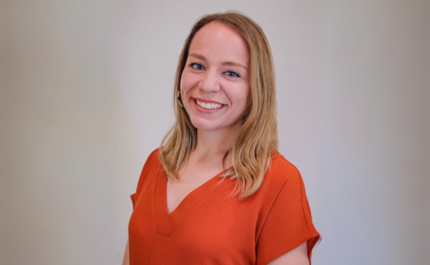 Francesca Genoese smiles for the camera while standing in front of a neutral gray background. Genoese wears a bright orange, v-neck tee shirt. Genoese has shoulder-length blonde hair.