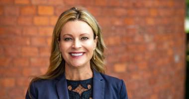 Tonya Frisbey headshot. Tonya poses in front of a reddish-orange brick building. Tonya wears a navy blue blazer over a black blouse. Tonya's blonde hair is curled and shoulder-length.
