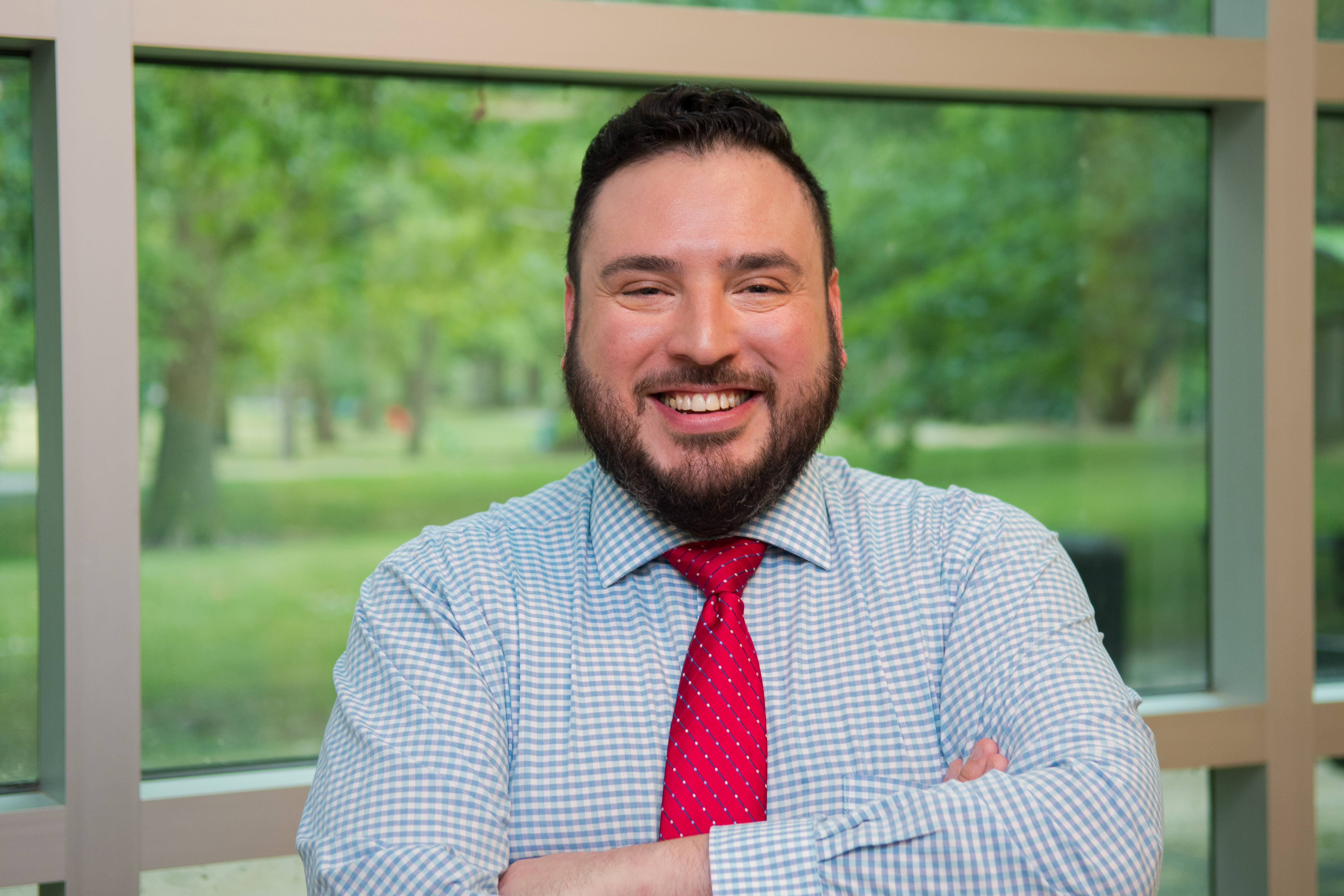 Gabriel smiles for the camera while crossing arms in front of the chest. Gabriel stands in front of large, rectangular glass windows overlooking a courtyard. Gabriel wears a light blue shirt and a red tie. Gabriel has short brown hair and a short brown beard.