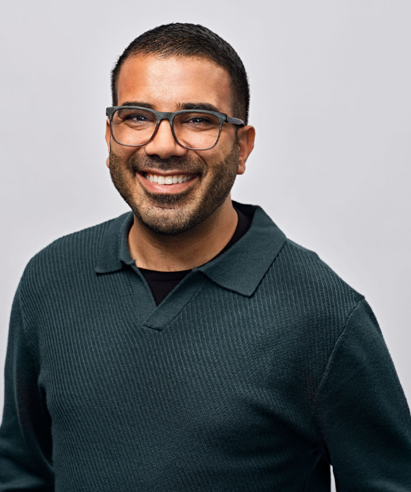 Omar Hakim smiles for a photo against a neutral gray backdrop. Hakim wears a dark green sweater and glasses with a black rim. Hakim's dark hair is cut close to the head.