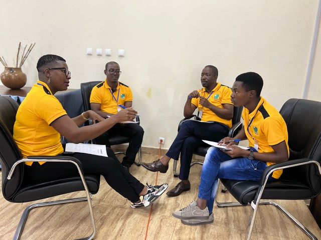 Four individuals wearing yellow and black polo shirts gather in a circle and discuss a topic. 