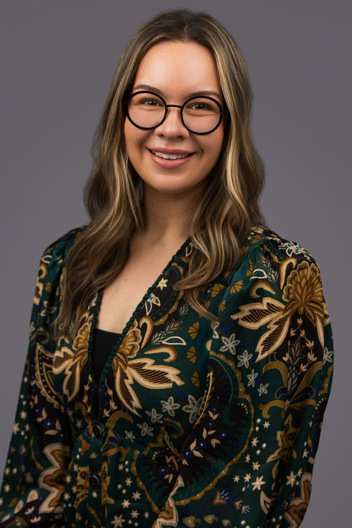 Pam Moura poses for the camera in front of a neutral gray background. Pam wears a black blazer covered in brown, beige and blue floral designs. Pam wears circular, dark-rimmed glasses.