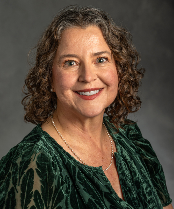 Mary Juzwik poses for the camera against a neutral gray background. Mary wears a dark green velvet top and a pearl necklace. Mary's dark hair is curly and about shoulder-length.