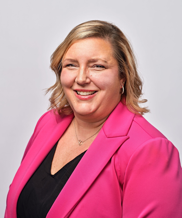 Venessa Keesler smiles for the camera against a neutral gray backdrop. Keesler wears a black blouse under a bright pink blazer. Keesler's hair is blonde, wavy and about shoulder-length.