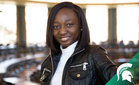 Olamide Ogungbemi headshot. Ogungbemi smiles in the kiva, a circular classroom in MSU's Erickson Hall. Ogungbemi wears a white shirt under a black leather jacket. Ogungbemi's black hair is shoulder-length.