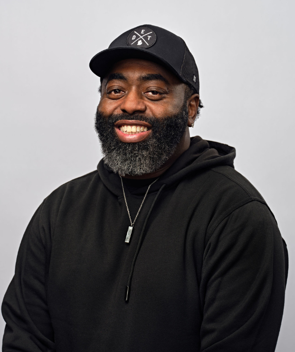 Curtis Lewis smiles for the camera against a neutral gray backdrop. Lewis wears a black hoodie and a black baseball cap with the word