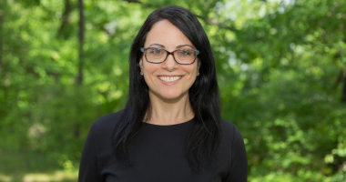 Joanne Marciano headshot. Joanne smiles toward the camera while standing outside in front of trees lush with green foliage. Joanne wears a black blouse and dark-rimmed glasses. Joanne's black hair falls past the shoulders.