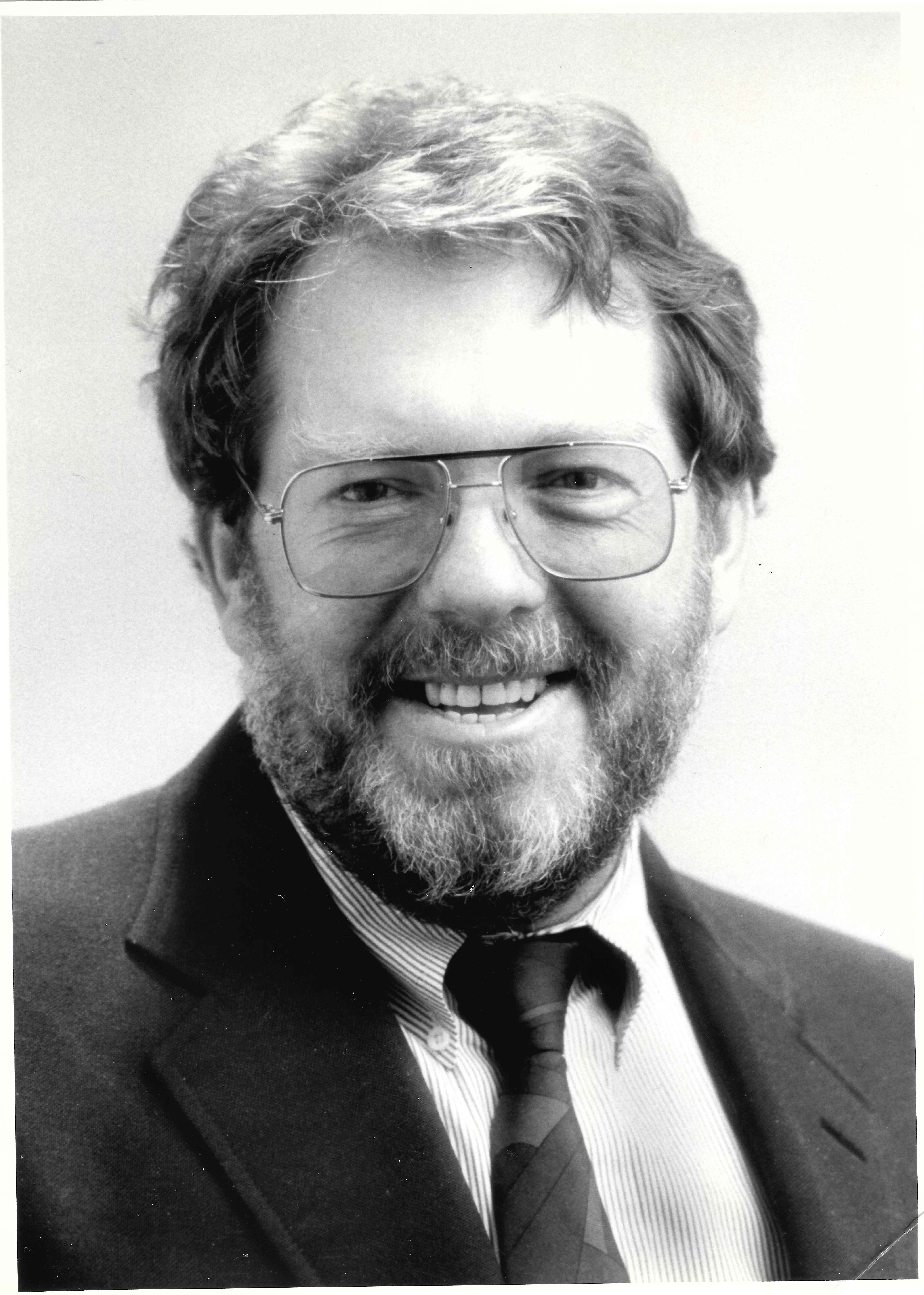 P. David Pearson smiles toward the camera against a neutral white background in this black and white photo. Pearson wears a dark suit and tie, a white button-down shirt and glasses. Pearson has a beard and short hair of a darkish color.