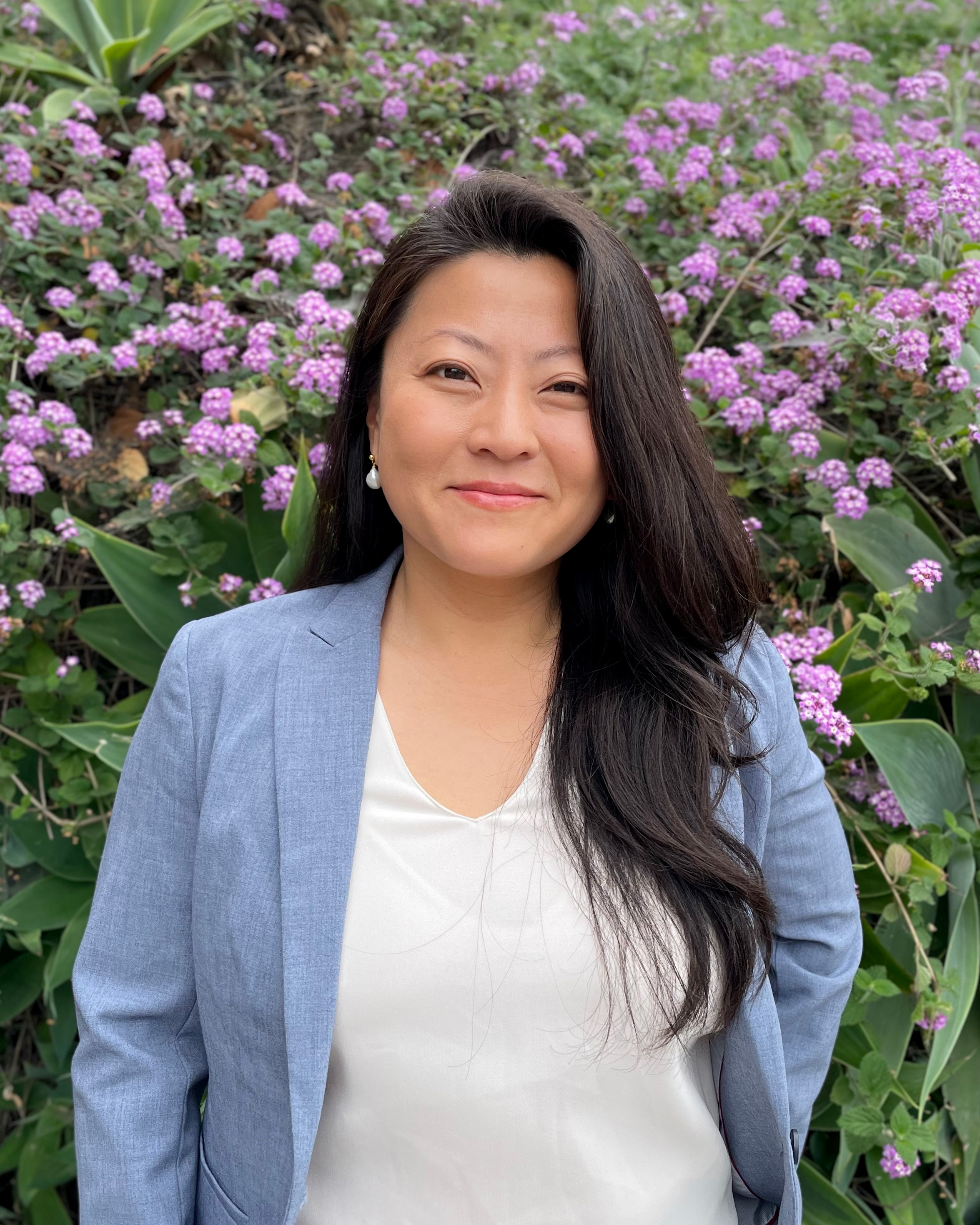 Sol smiles for the camera while standing in front of a bush lush with green leaves and purple flowers. Sol wears a white blouse under a light blue blazer. Sol's hair is long and black. 