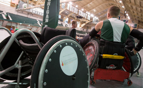 Close-up of adaptive wheelchairs used in Sport settings. The wheels are dark with silver-colored metal frames. An individual wearing a green and white jersey leans away from the camera.