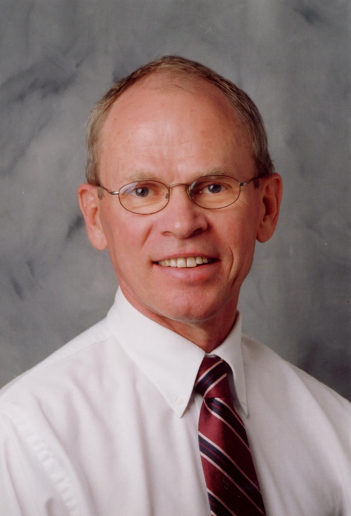 Jack smiles for the camera against a neutral gray background. Jack has a white button-down shirt and a red and black patterned tie.