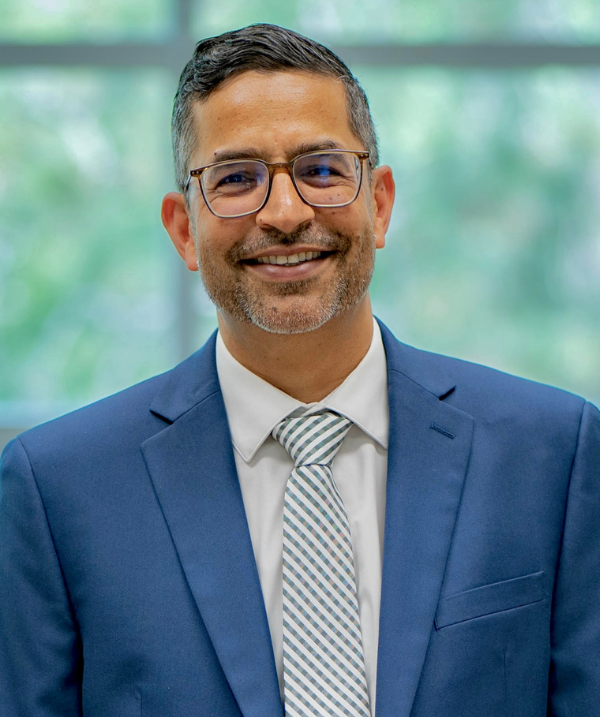 Aman Yadav smiles for the camera while standing in front of Erickson Hall's horizontal rectangle windows. Yadav wears a white button-down shirt, and a green and white striped tie with a blue suit jacket. Yadav also has dark-rimmed glasses on. Yadav has black and gray short hair and a slight beard.
