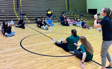 Corinne Zimmerman talks into a microphone and talks to a group of children playing in various sports along a gymnasium floor.
