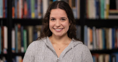 Madison Pizzuti headshot. Madison poses against a full bookshelf. Madison wears a gray sweater. Madison has long, brown hair.