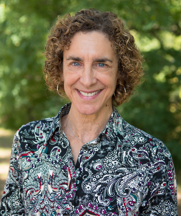 Gail Richmond smiles outside. Richmond stands in front of trees lush with green foliage. Richmond wears a shirt with a black, red, green and white paisley pattern. Richmond has silver-hooped earrings on. Richmond's brown hair is curly and about chin-length.