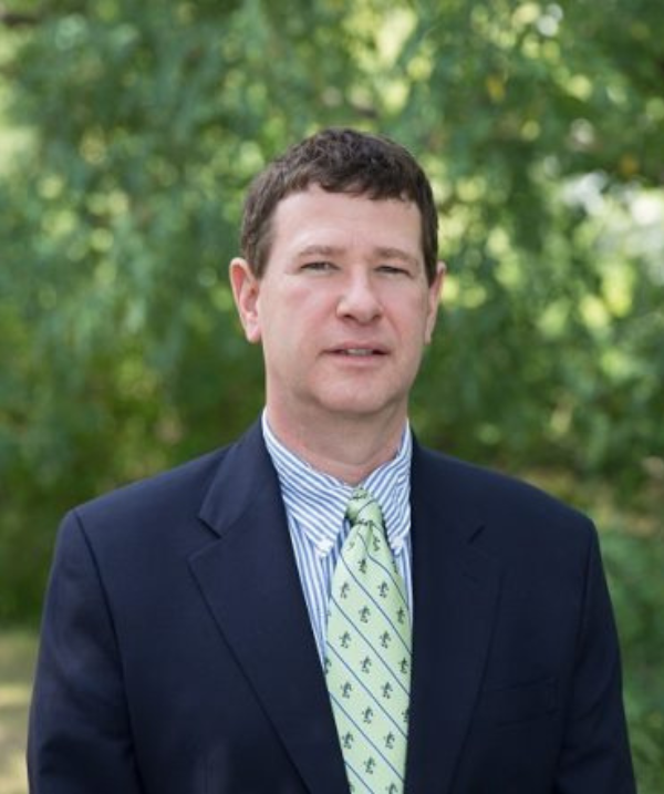 Matthew Warzynski smiles for the camera in an outdoor setting, standing in front of bushes lush with green leaves. Matthew wears a blue and white stripped button down shirt and a light green tie as well as a navy blue blazer. Matthew's brown hair is short.