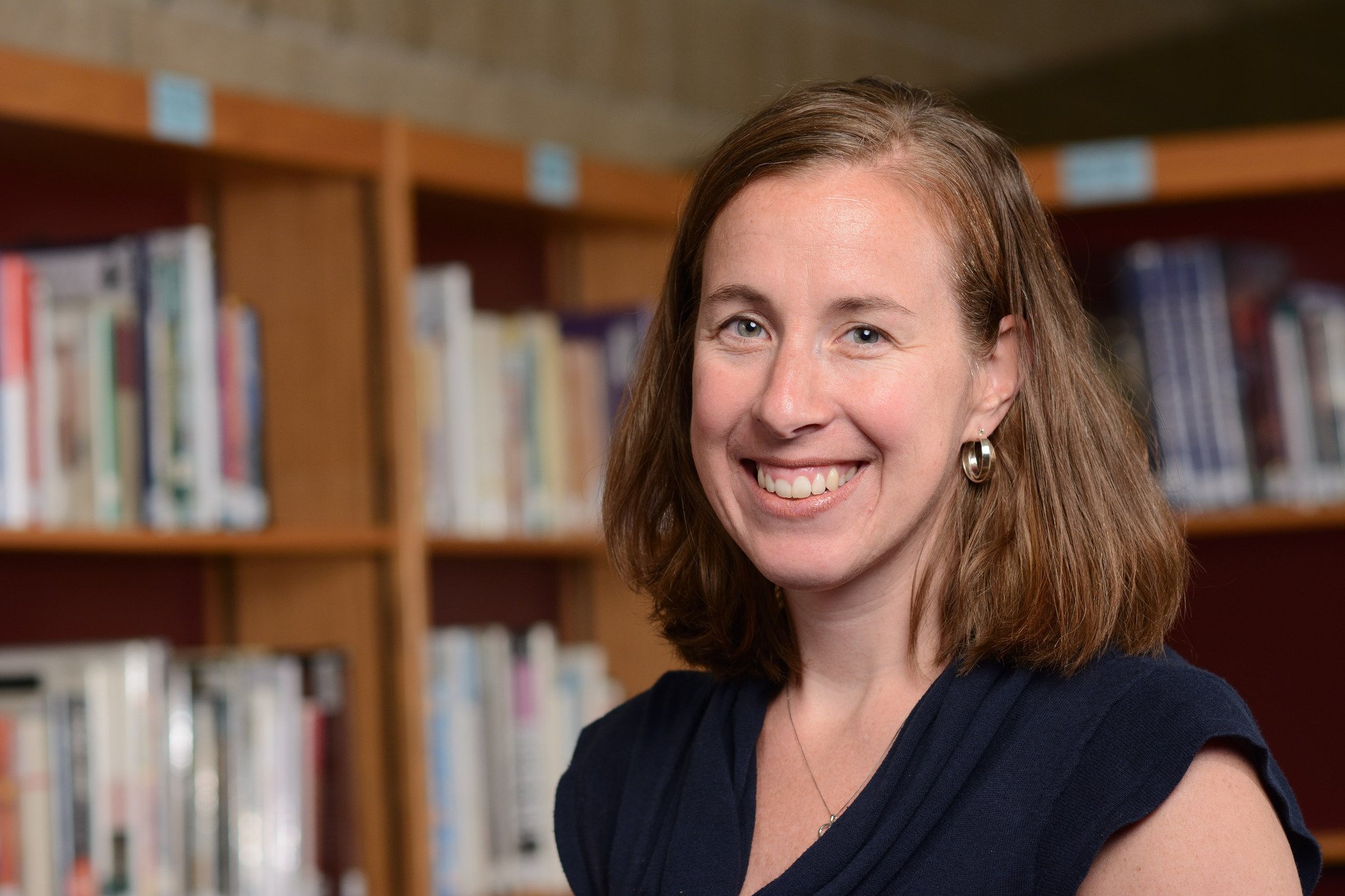 Tara Becker-Utess headshot. Becker-Utess poses in front of a bookshelf. Becker-Utess wears a black shirt. 