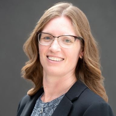 Sarah Witmer smiles for the camera against a neutral gray background. Sarah wears a gray top under a black blazer, glasses with dark upper rims, and a necklace with a silver pendant. Sarah's reddish hair falls past the shoulders in waves.