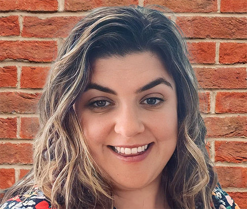 mallory with a brick background and long wavy brown hair with blond highlights. she is wearing a patterned blouse.