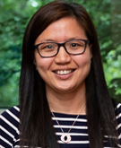 Connie Sung headshot. Connie smiles toward the camera while standing in front of a bush lush with green leaves. Connie wears a black and white horizontally striped shirt and black-rimmed glasses. Connie's black hair is long and falls past the shoulders.