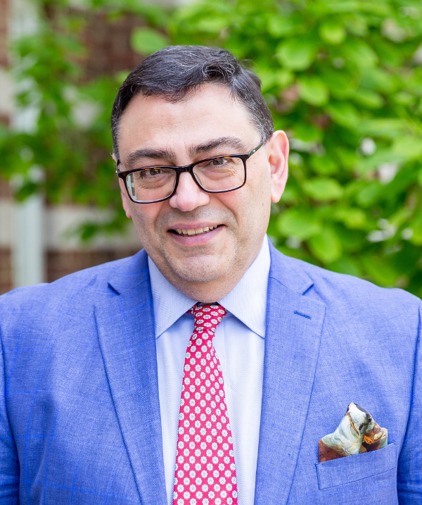 Paddy Ekkekakis smiles for the camera in front of a bush lush with green leaves. Ekkekakis wears a white button-down shirt, a red tie and a blue blazer. Ekkekakis has dark-rimmed glasses and black hair cut close to the head