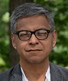 Higinio Dominguez poses for the camera while standing in an outdoor location. Dominguez wears a white button-down shirt under a gray blazer and dark-rimmed glasses. Dominguez has black and gray short hair.