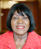 Patricia Edwards smiles for the camera while standing in a hallway. Edwards wears a bright red shirt. Edwards has black, shoulder-length hair and bangs.