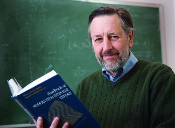 Mark Reckase smiles toward the camera while positioned in front of a traditional green chalkboard. Reckase holds up a book. Reckase has dark brown and gray hair and a short beard