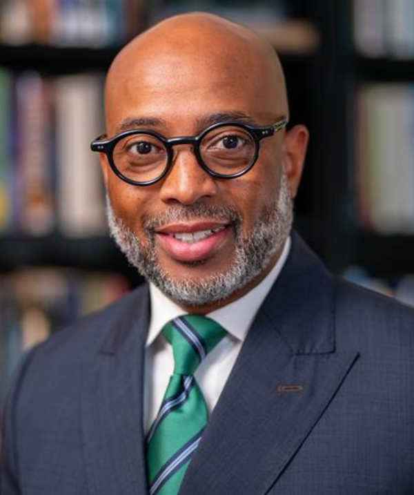 Jerlando Jackson headshot Jerlando Jackson headshot. Jackson poses in front of a bookshelf. Jackson wears a dark blazer over a green tie. Jackson has dark-rimmed glasses.