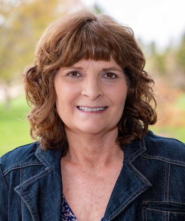 Michelle Hatta smiles for the camera in an outdoor setting. Michelle wears a dark denim jacket over a floral blouse. Michelle's brown hair is curly; Michelle has bangs.
