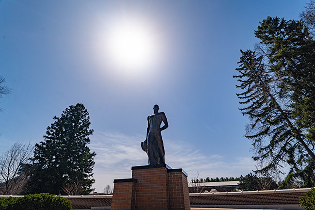 The Spartan statue on the Michigan State University campus, standing prominently against a clear blue sky with the bright sun shining above. The scene is framed by tall evergreen trees and brick structures in the background.