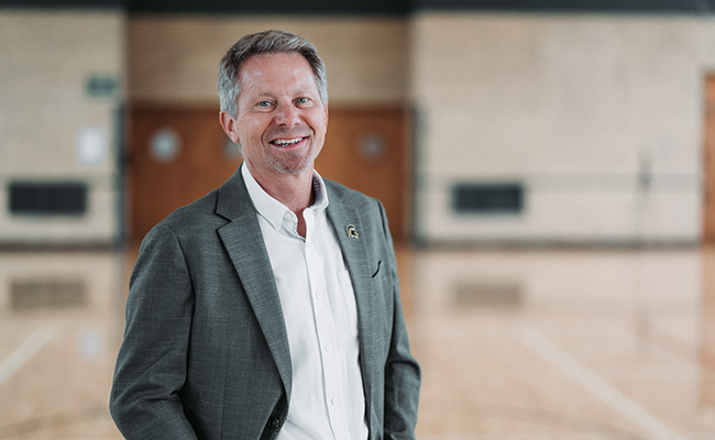A smiling individual with short gray hair, wearing a gray blazer, white shirt, and a lapel pin, standing indoors in what appears to be a gymnasium or multipurpose space with wooden floors and blurred doors in the background.