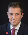 A professional portrait of an individual with short, neatly styled hair, wearing a dark suit, white shirt, and red tie, with a red lapel pin, set against a dark background.