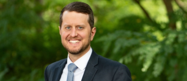 A professional headshot of Matt Harkey. He is standing outdoors and wearing a suit.
