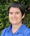 A smiling individual with short dark hair, wearing a blue shirt, standing outdoors with greenery in the background.