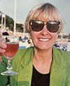 Laura Roehler-Carmean smiles for the camera while in a beachy setting. Roehler-Carmean holds up a goblet with a reddish liquid. Roehler-Carmean wears a black top under a bright green blazer and black-rimmed sunglasses. Roehler-Carmean's blonde hair is short, with bangs falling slightly over the sunglasses.