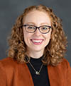 An individual with curly red hair and glasses, wearing a rust-colored blazer over a black top with a necklace, set against a dark gray background.
