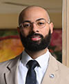 An individual with a beard and glasses, wearing a beige suit, white shirt, and black tie, standing in front of a warm-toned background.
